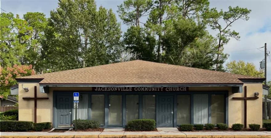 Photograph of the single-storey Jacksonville Community Church building, its name lettered above a row of glass doors, a wooden cross at each end and an accessible parking space beside the entrance
