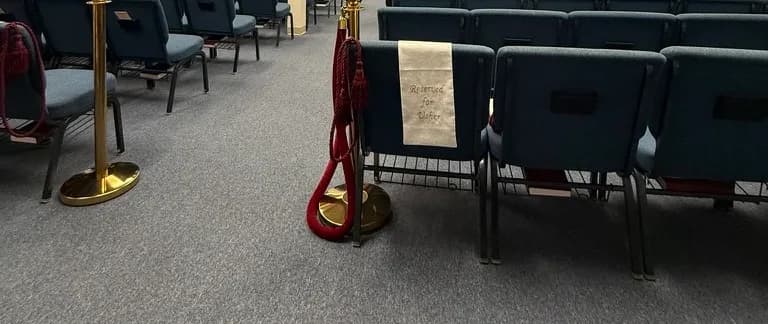 Photograph of the sanctuary's empty teal chairs beside a brass stanchion hung with a red velvet rope, a cloth marked 'Reserved for Usher' draped over one seat back